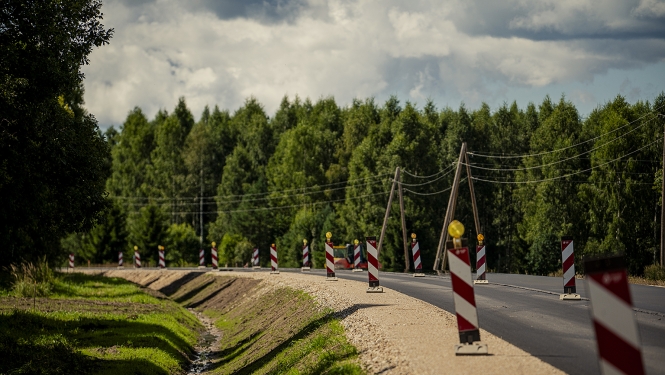 Līkums uz asfaltēta ceļa meža apvidū. Ceļa malā izvietoti sarkanbalti ceļa remontdarbu norobežojošie stabiņi, bet fonā redzamas augstas egles un mākoņainas debesis. Blakus ceļam ir grants mala un grāvis.
