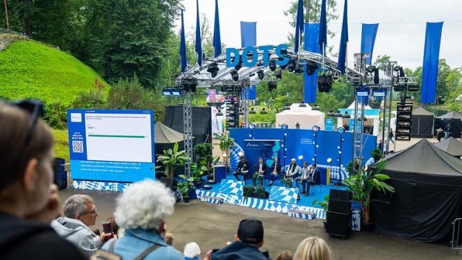 Sarunu festivāla LAMPA skatuvē “DOTS” notiek diskusija par Rail Baltica un Rīgas Centrālo staciju. Uz skatuves sēž diskusijas dalībnieki, bet skatītāji klausās no amfiteātrim līdzīgām sēdvietām. Blakus skatuvei redzams ekrāns ar balsojuma rezultātiem. Skatuvi rotā zilas dekorācijas un Rail Baltica informatīvie materiāli. Fonā redzama festivāla teritorija un zaļā apkārtne Cēsīs.