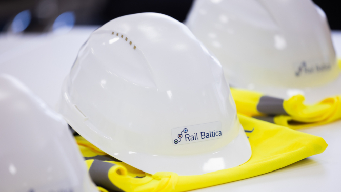 Uz galda novietoti balti aizsargķiveres ar uzlīmi 'Rail Baltica' un dzelteni atstarojoši vestes fragmenti