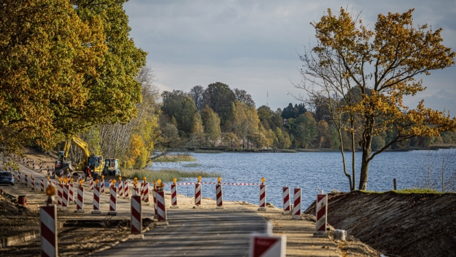 Rudens ainava pie ūdens: koka laipa ar sarkanbaltiem ceļa norobežojumiem ved uz ezeru vai upi, krastā aug koki ar dzeltenām un zaļām lapām, bet fonā redzami koki un mierīgs ūdens.