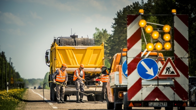 Valsts autoceļa remontdarbi. Uz ceļa strādā vairāki ceļu būves darbinieki augstas redzamības vestēs. Blakus atrodas dzeltena kravas automašīna un ceļu uzturēšanas transportlīdzeklis ar mirgojošām brīdinājuma gaismām un ceļa darbu zīmēm. Darbi notiek uz brauktuves meža ieskautā teritorijā saulainā dienā.