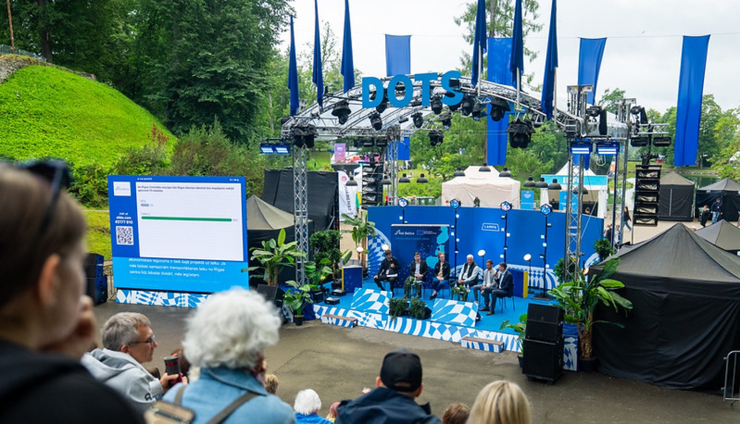 Sarunu festivāla LAMPA skatuvē “DOTS” notiek diskusija par Rail Baltica un Rīgas Centrālo staciju. Uz skatuves sēž diskusijas dalībnieki, bet skatītāji klausās no amfiteātrim līdzīgām sēdvietām. Blakus skatuvei redzams ekrāns ar balsojuma rezultātiem. Skatuvi rotā zilas dekorācijas un Rail Baltica informatīvie materiāli. Fonā redzama festivāla teritorija un zaļā apkārtne Cēsīs.