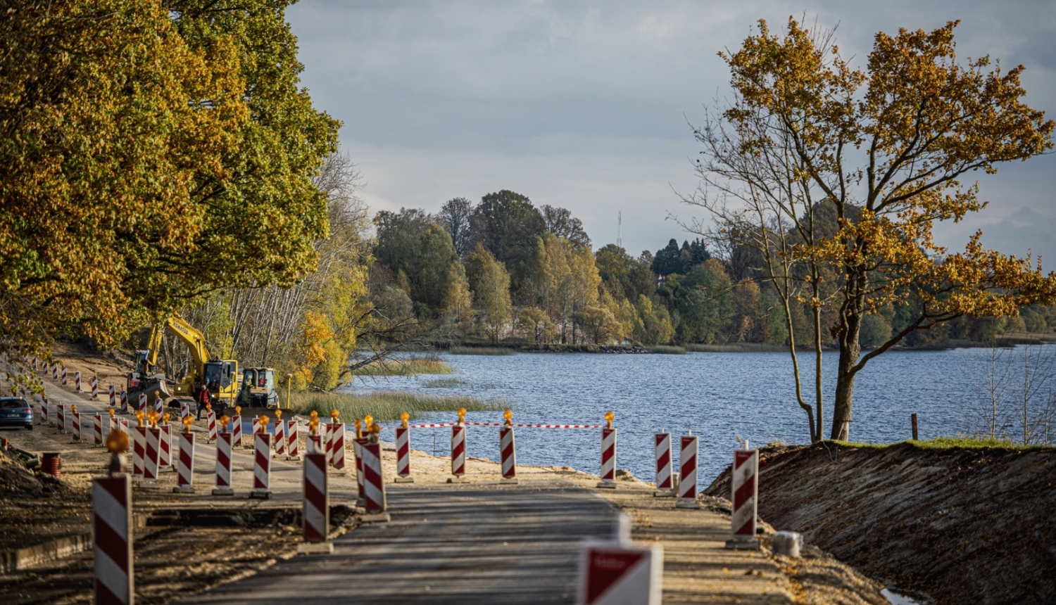 Rudens ainava pie ūdens: koka laipa ar sarkanbaltiem ceļa norobežojumiem ved uz ezeru vai upi, krastā aug koki ar dzeltenām un zaļām lapām, bet fonā redzami koki un mierīgs ūdens.