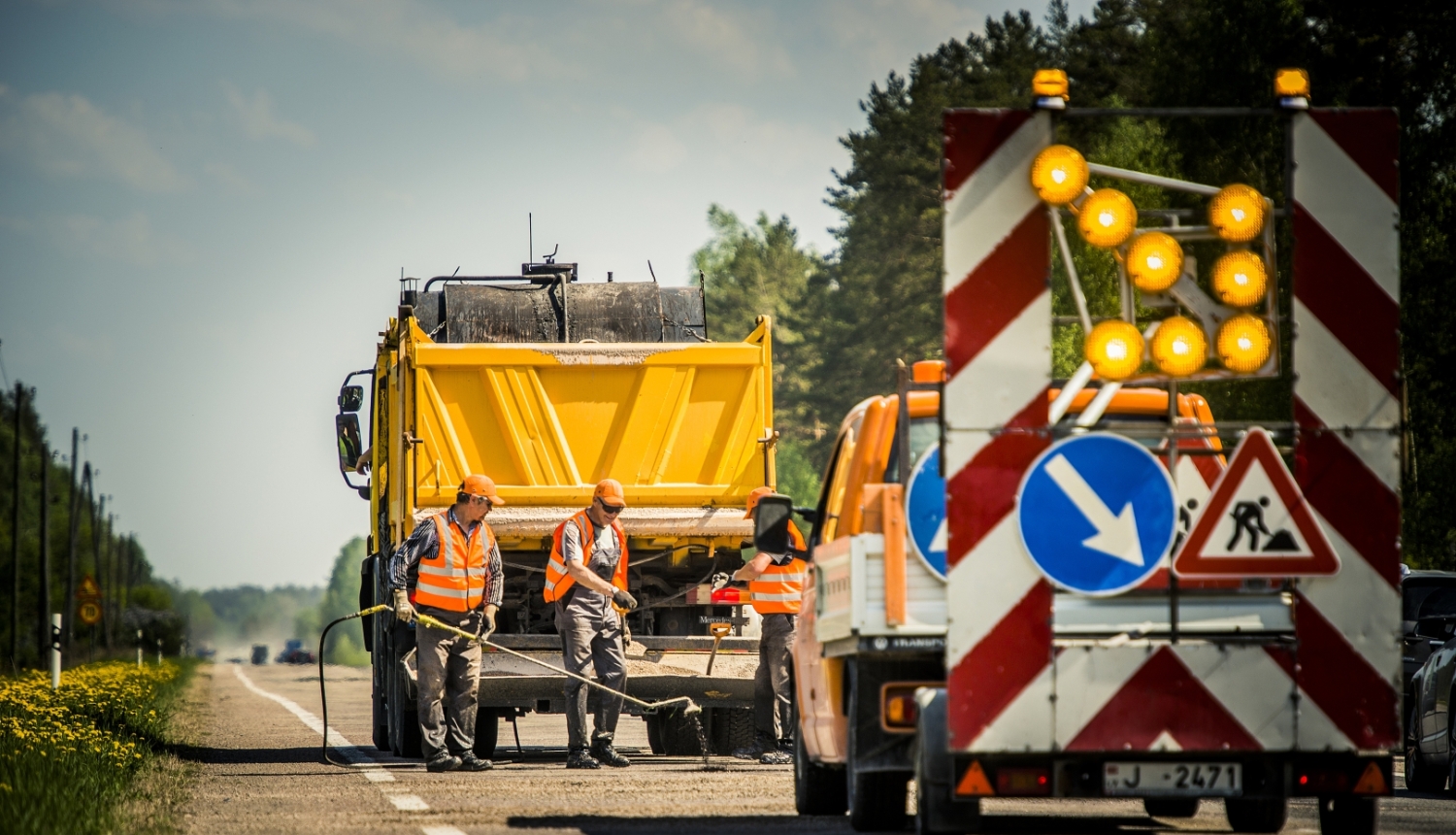Valsts autoceļa remontdarbi. Uz ceļa strādā vairāki ceļu būves darbinieki augstas redzamības vestēs. Blakus atrodas dzeltena kravas automašīna un ceļu uzturēšanas transportlīdzeklis ar mirgojošām brīdinājuma gaismām un ceļa darbu zīmēm. Darbi notiek uz brauktuves meža ieskautā teritorijā saulainā dienā.