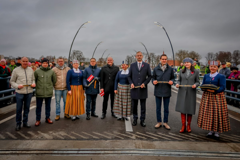 Salacas tilta atklāšanas pasākums Salacgrīvā. Uz tilta rindā stāv cilvēku grupa, daļa tērpušies tautas tērpos, citi – mēteļos. Viņi tur lentas un Latvijas karogu, gatavojoties svinīgai lentas griešanas ceremonijai. Fonā redzami skatītāji un tilta margas