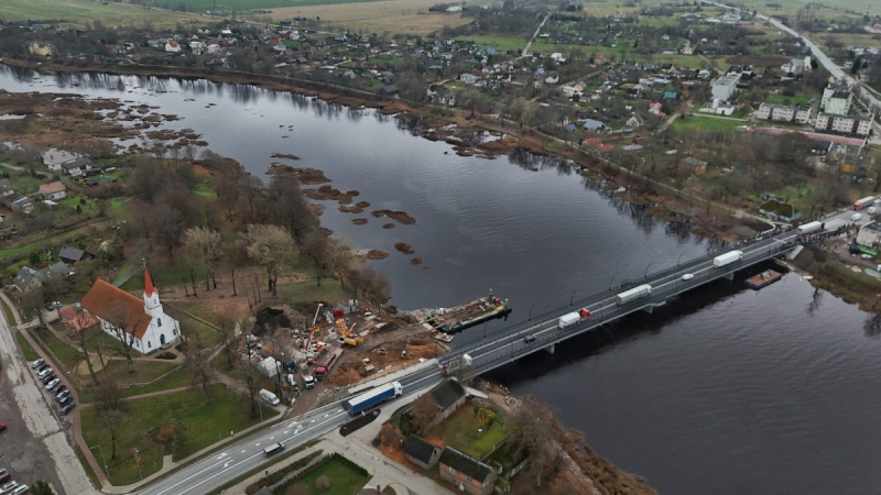 Salacas tilts Salacgrīvā, skatīts no augšas. Tilts šķērso upi, uz tā redzami vairāki transportlīdzekļi. Kreisajā pusē pie tilta atrodas balta baznīca ar sarkanu jumtu un apkārt koki, blakus redzama būvlaukuma tehnika. Fonā redzama pilsētas apbūve un lauki