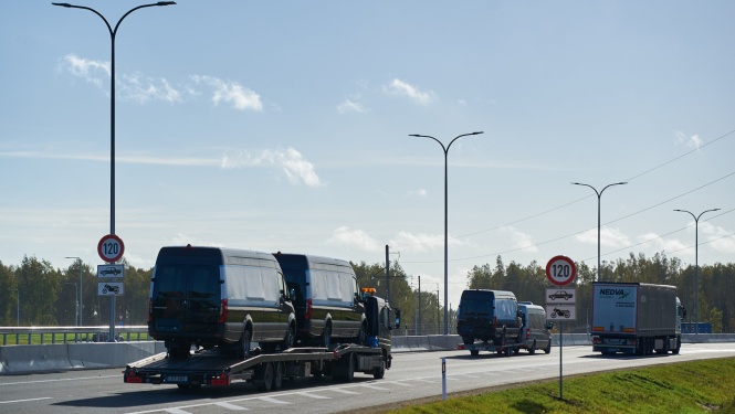 Kravas automašīna ved divus melnus mikroautobusus uz platformas pa jaunu ceļa posmu ar atļauto ātrumu 120 km/h. Blakus brauc citi transportlīdzekļi, redzamas ceļa zīmes un apgaismes stabi, fonā koki un skaidras debesis