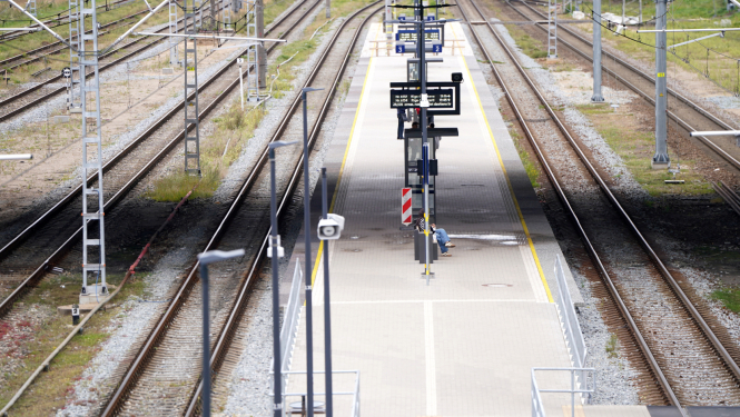 Tukša dzelzceļa stacija ar vairākām sliežu joslām un centrālo peronu. Uz perona redzamas informācijas tabulas, apgaismojuma stabi un viena persona, kas strādā pie aprīkojuma. Fonā redzamas elektrības līnijas un sliežu infrastruktūra
