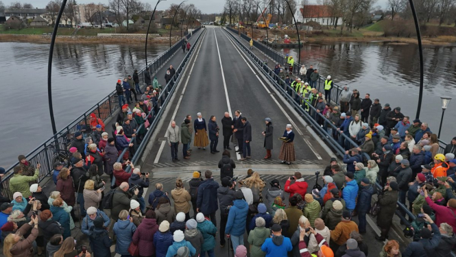 Salacas tilta atklāšanas pasākums Salacgrīvā. Uz tilta stāv cilvēku grupa, kas piedalās ceremonijā, bet abās pusēs gar margām un uz tilta sākuma ir sapulcējušies daudzi skatītāji. Tilts šķērso upi, fonā redzamas mājas, koki un celtniecības tehnika