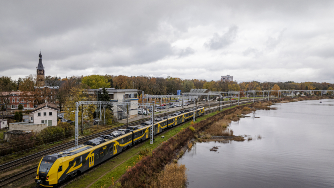 Drona foto ar Latvijas ainavu, kur vienā pusē ir ūdens, otrā pusē - pilsēta un pa vidu sliedes, pa kurām brauc Vivi vilciens