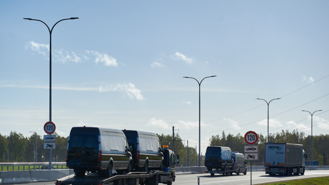 Kravas automašīna ved divus melnus mikroautobusus uz platformas pa jaunu ceļa posmu ar atļauto ātrumu 120 km/h. Blakus brauc citi transportlīdzekļi, redzamas ceļa zīmes un apgaismes stabi, fonā koki un skaidras debesis