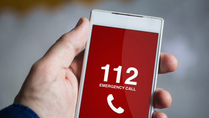 Cilvēka roka tur mobilo telefonu. Telefona displejā redzams telefona numurs 112