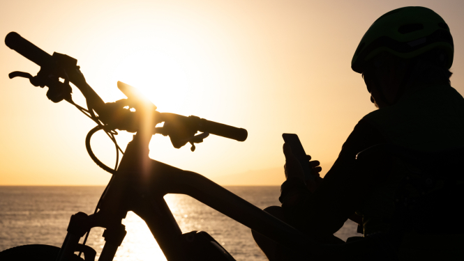 Cilvēks ar velosipēdu sēž pie jūras saulrietā un skatās telefonā. Redzams tikai cilvēka un velosipēda siluets