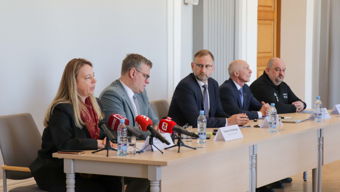 Preses konferences pieci dalībnieki sēž pie galdiem uz kuriem ir mediju mikrofoni