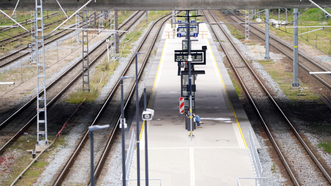 Tukša dzelzceļa stacija ar vairākām sliežu joslām un centrālo peronu. Uz perona redzamas informācijas tabulas, apgaismojuma stabi un viena persona, kas strādā pie aprīkojuma. Fonā redzamas elektrības līnijas un sliežu infrastruktūra