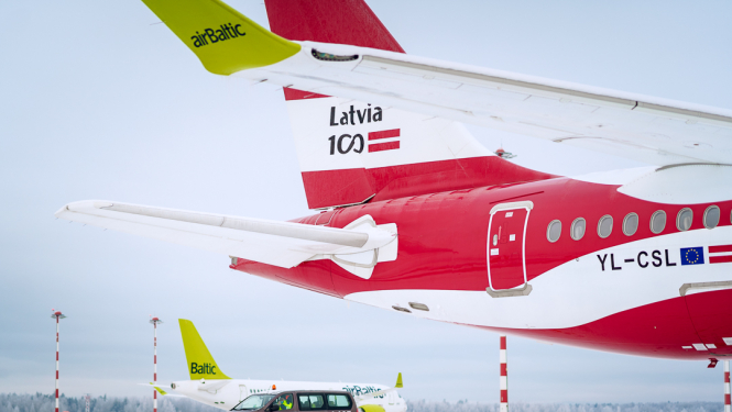Lidostas skrejceļš ziemas apstākļos ar sniegu. Priekšplānā redzama airBaltic lidmašīna ar sarkanu korpusu un uzrakstu 'Latvia 100' uz astes, kā arī reģistrācijas numuru YL-CSL. Blakus lidmašīnai stāv brūns lidostas servisa mikroautobuss ar numuru L125. Fonā redzamas vēl divas airBaltic lidmašīnas ar zaļām astēm un sarkanbaltiem gaismas stabiem. Debesis ir pelēkas, fonā mežs ar apsnigušiem kokiem