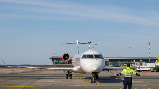 Lidlauka peronā redzama lidmašīna, kas lēni tuvojas stāvvietai. Lidmašīnas priekšā stāv lidostas darbinieks ar drošības vesti un tur signālvālītes, ar kurām norāda lidmašīnai kustības virzienu. Fonā redzama lidostas ēka, skrejceļi un aprīkojums.