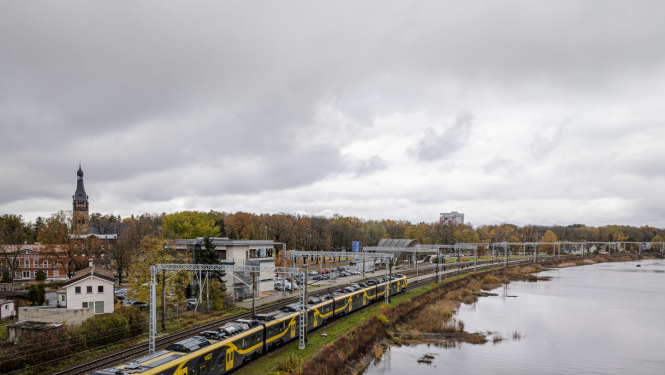 Drona foto ar Latvijas ainavu, kur vienā pusē ir ūdens, otrā pusē - pilsēta un pa vidu sliedes, pa kurām brauc Vivi vilciens
