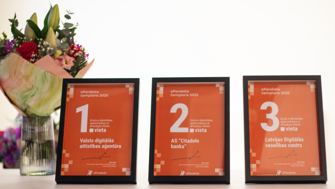Uz galda rindā novietoti trīs ierāmēti apbalvojumi “eParaksta čempions 2025”: 1. vieta – Valsts digitālās attīstības aģentūra, 2. vieta – AS “Citadele banka”, 3. vieta – Latvijas Digitālais veselības centrs. Kreisajā pusē redzams ziedu pušķis vāzē.
