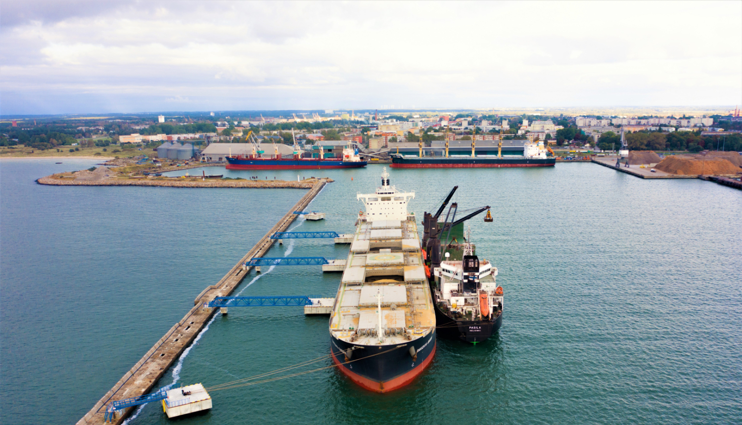Skats no augšas uz jūras ostu: divi lieli kravas kuģi piestātnē, blakus darba kuģis ar celtni. Priekšplānā mols un pontoni, fonā pilsēta un rūpniecības teritorija piekrastē.