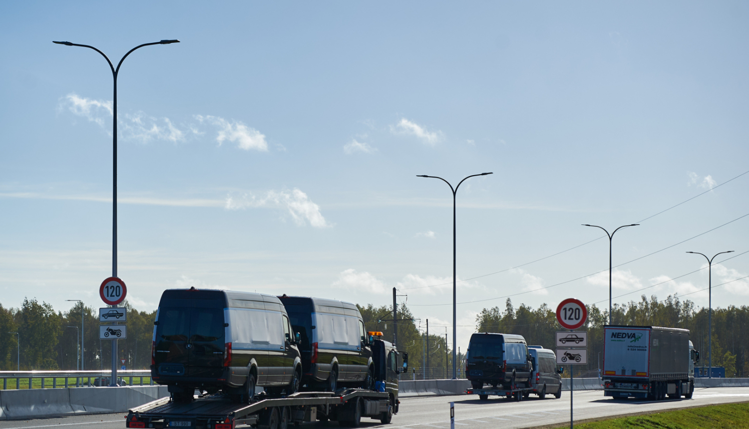 Kravas automašīna ved divus melnus mikroautobusus uz platformas pa jaunu ceļa posmu ar atļauto ātrumu 120 km/h. Blakus brauc citi transportlīdzekļi, redzamas ceļa zīmes un apgaismes stabi, fonā koki un skaidras debesis