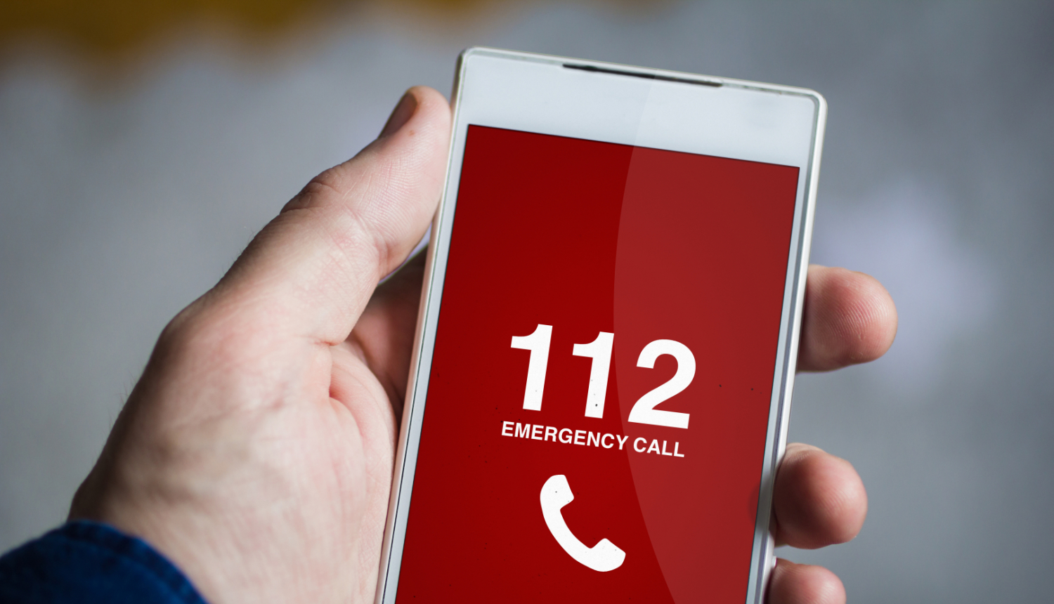 Cilvēka roka tur mobilo telefonu. Telefona displejā redzams telefona numurs 112