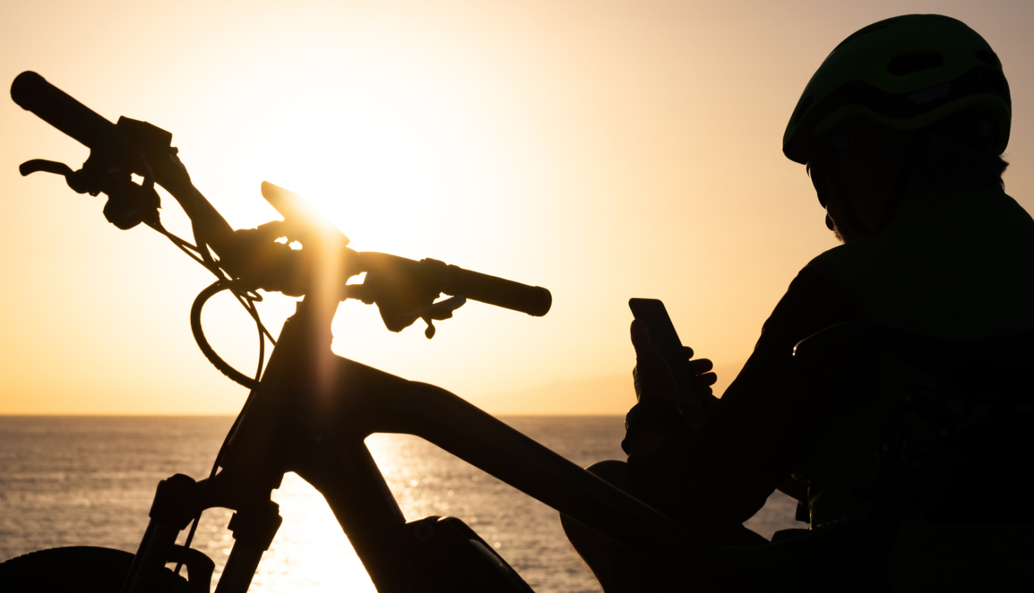 Cilvēks ar velosipēdu sēž pie jūras saulrietā un skatās telefonā. Redzams tikai cilvēka un velosipēda siluets