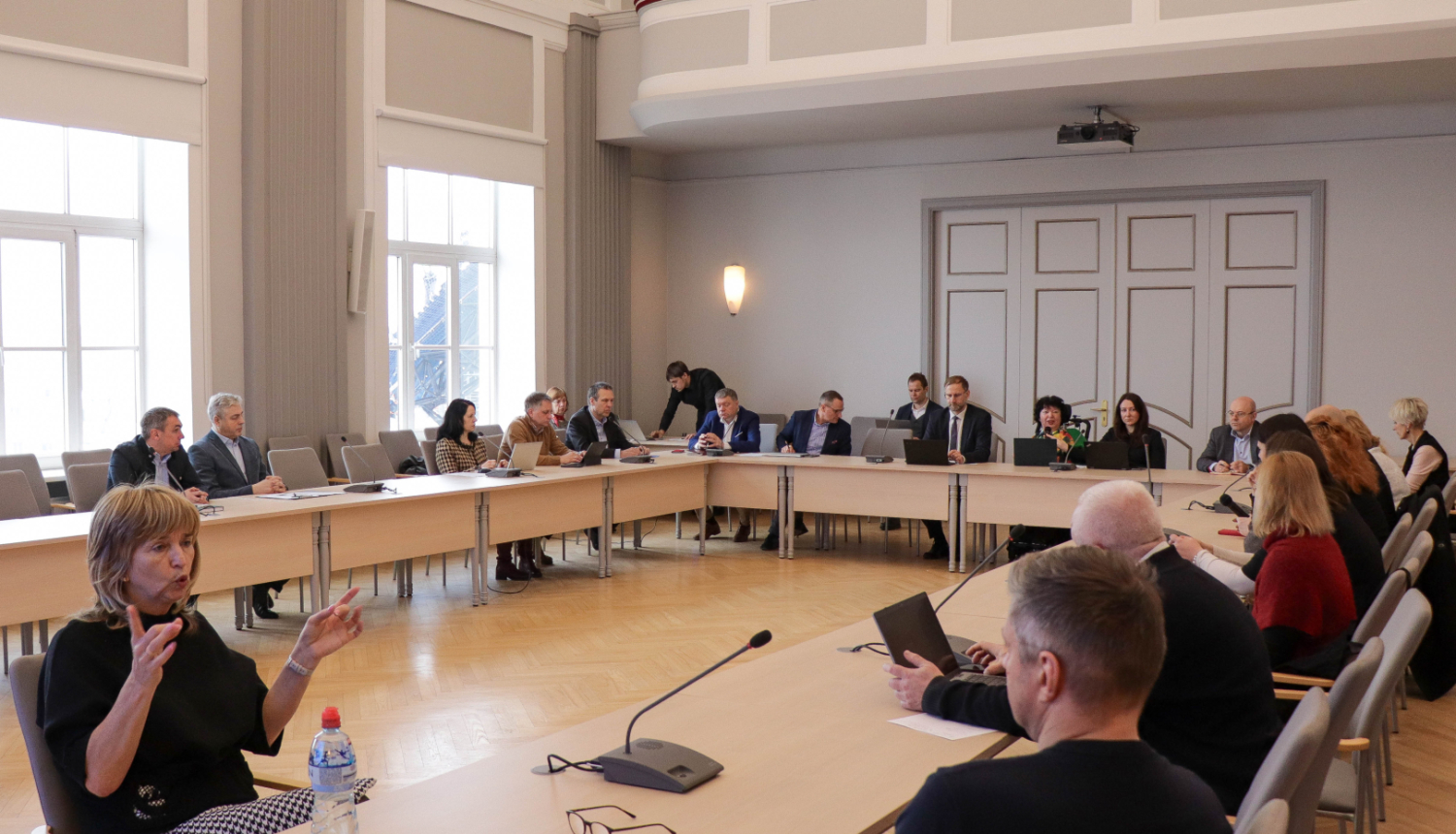Mobilitātes padomes sēde ministrijas lielajā zālē. Visi dalībnieki sēž pie galdiem un diskutē