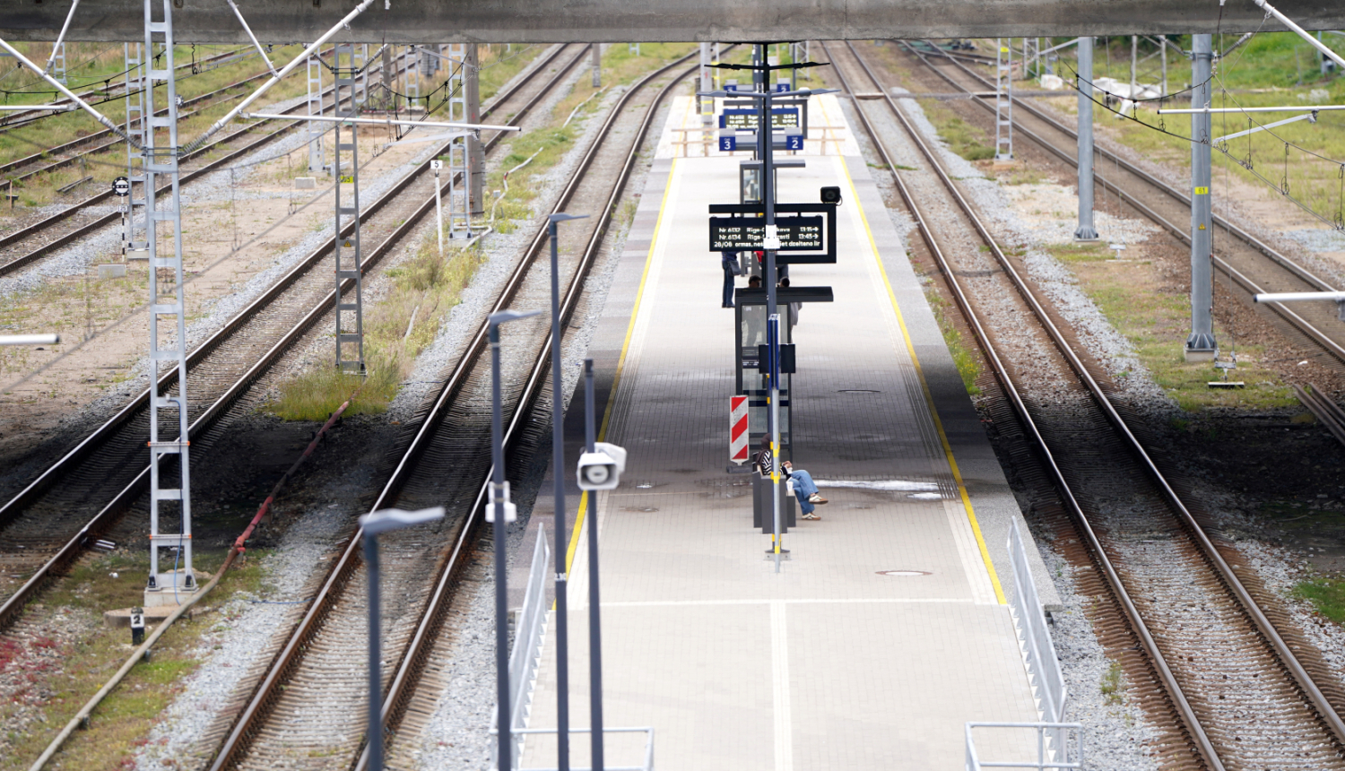 Tukša dzelzceļa stacija ar vairākām sliežu joslām un centrālo peronu. Uz perona redzamas informācijas tabulas, apgaismojuma stabi un viena persona, kas strādā pie aprīkojuma. Fonā redzamas elektrības līnijas un sliežu infrastruktūra