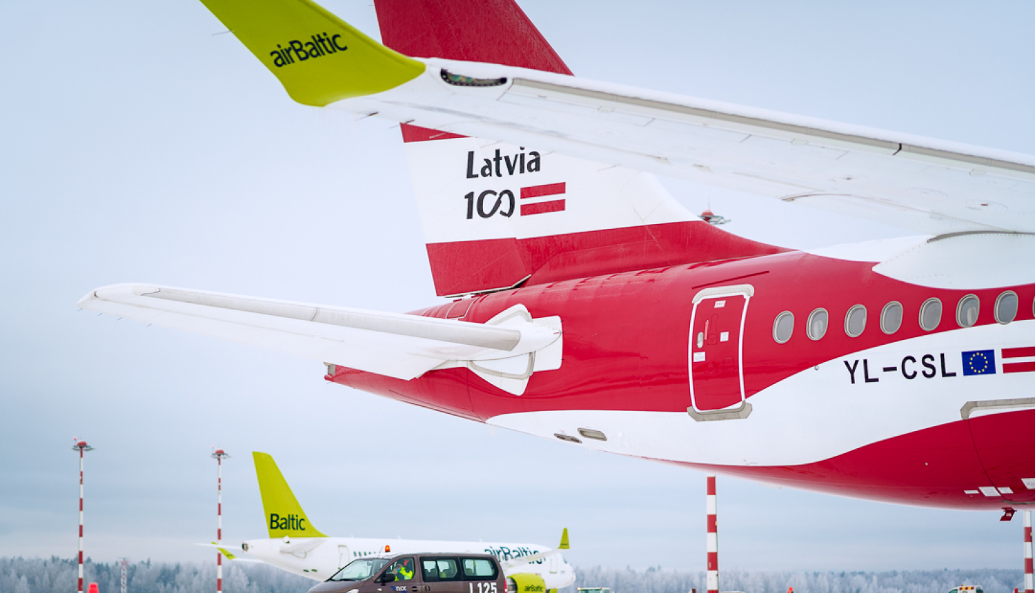 Lidostas skrejceļš ziemas apstākļos ar sniegu. Priekšplānā redzama airBaltic lidmašīna ar sarkanu korpusu un uzrakstu 'Latvia 100' uz astes, kā arī reģistrācijas numuru YL-CSL. Blakus lidmašīnai stāv brūns lidostas servisa mikroautobuss ar numuru L125. Fonā redzamas vēl divas airBaltic lidmašīnas ar zaļām astēm un sarkanbaltiem gaismas stabiem. Debesis ir pelēkas, fonā mežs ar apsnigušiem kokiem