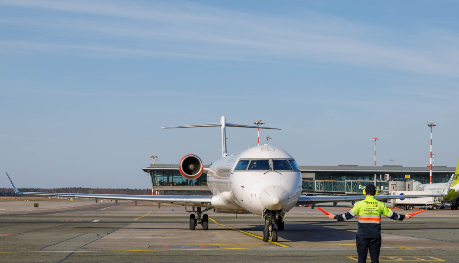 Lidlauka peronā redzama lidmašīna, kas lēni tuvojas stāvvietai. Lidmašīnas priekšā stāv lidostas darbinieks ar drošības vesti un tur signālvālītes, ar kurām norāda lidmašīnai kustības virzienu. Fonā redzama lidostas ēka, skrejceļi un aprīkojums.