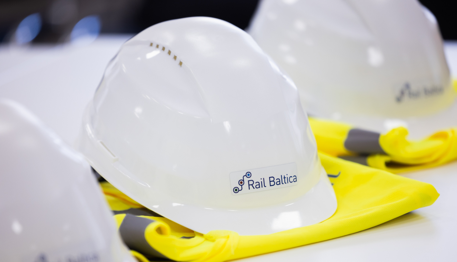 Uz galda novietoti balti aizsargķiveres ar uzlīmi 'Rail Baltica' un dzelteni atstarojoši vestes fragmenti
