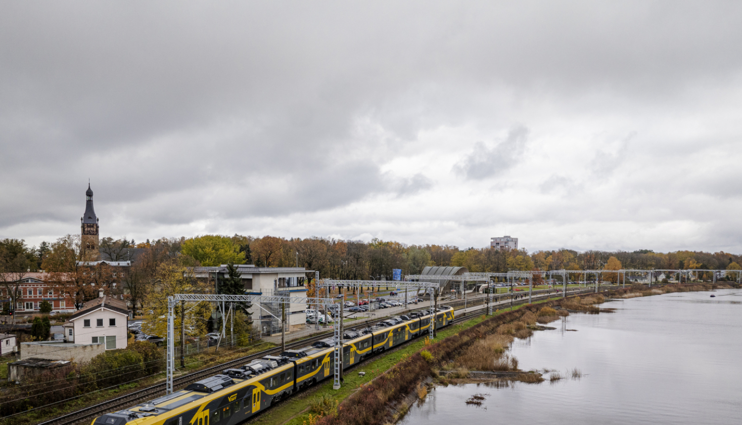 Drona foto ar Latvijas ainavu, kur vienā pusē ir ūdens, otrā pusē - pilsēta un pa vidu sliedes, pa kurām brauc Vivi vilciens