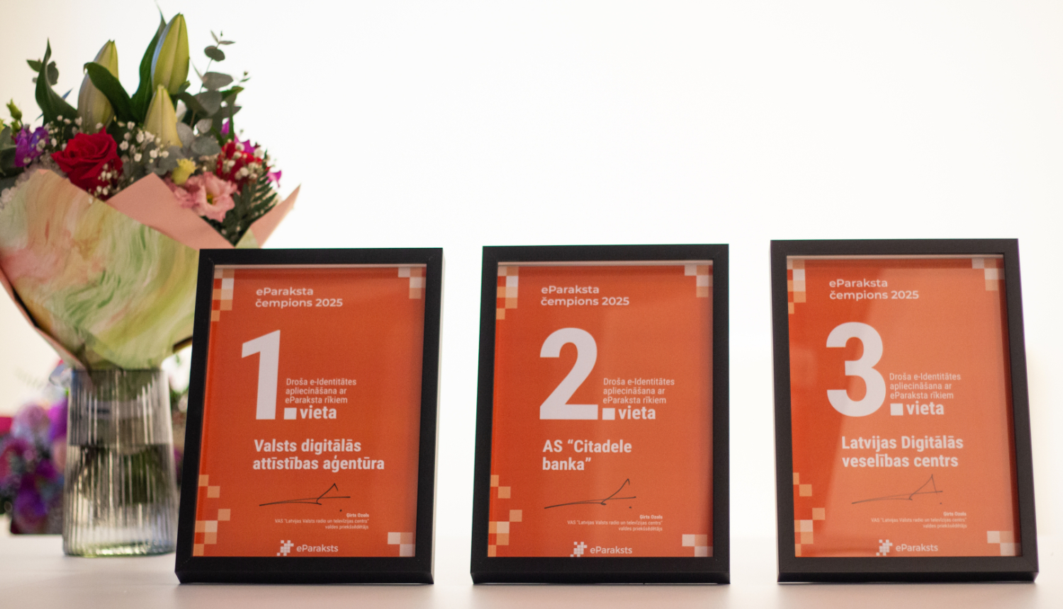 Uz galda rindā novietoti trīs ierāmēti apbalvojumi “eParaksta čempions 2025”: 1. vieta – Valsts digitālās attīstības aģentūra, 2. vieta – AS “Citadele banka”, 3. vieta – Latvijas Digitālais veselības centrs. Kreisajā pusē redzams ziedu pušķis vāzē.