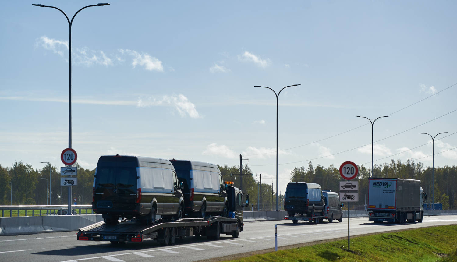 Kravas automašīna ved divus melnus mikroautobusus uz platformas pa jaunu ceļa posmu ar atļauto ātrumu 120 km/h. Blakus brauc citi transportlīdzekļi, redzamas ceļa zīmes un apgaismes stabi, fonā koki un skaidras debesis