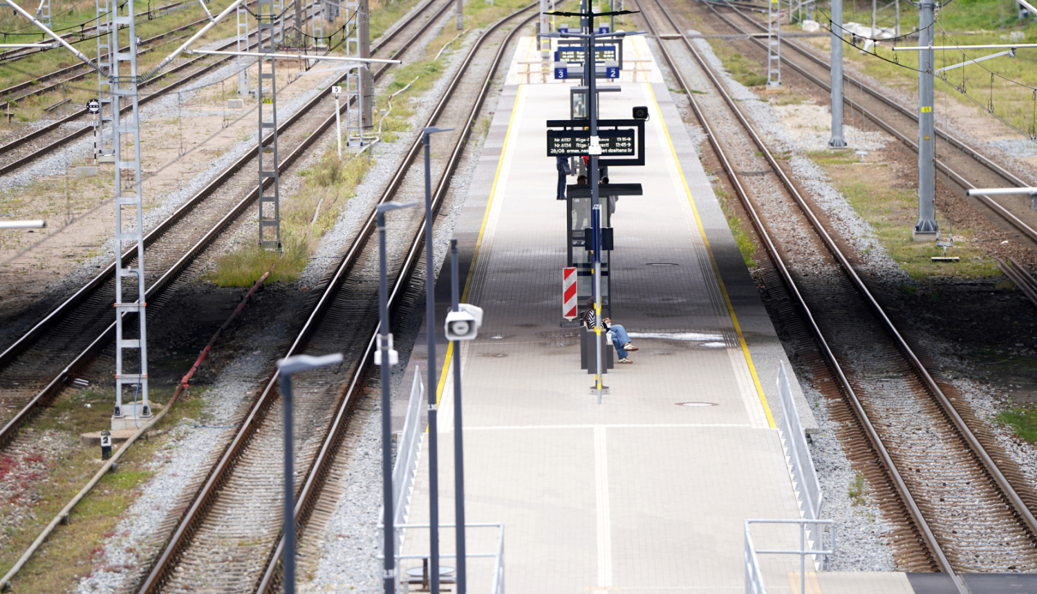 Tukša dzelzceļa stacija ar vairākām sliežu joslām un centrālo peronu. Uz perona redzamas informācijas tabulas, apgaismojuma stabi un viena persona, kas strādā pie aprīkojuma. Fonā redzamas elektrības līnijas un sliežu infrastruktūra