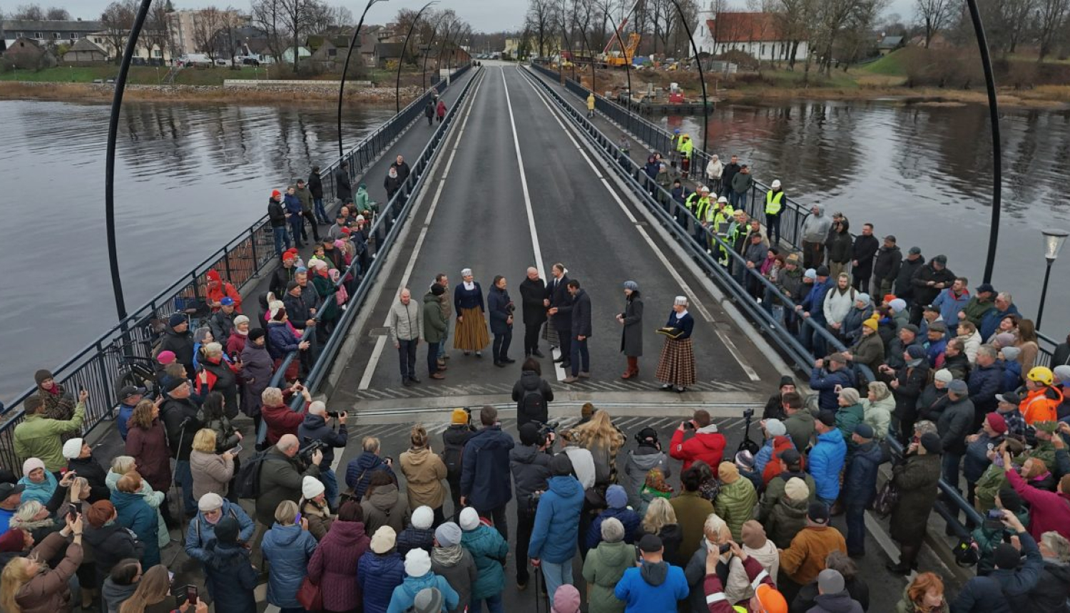 Salacas tilta atklāšanas pasākums Salacgrīvā. Uz tilta stāv cilvēku grupa, kas piedalās ceremonijā, bet abās pusēs gar margām un uz tilta sākuma ir sapulcējušies daudzi skatītāji. Tilts šķērso upi, fonā redzamas mājas, koki un celtniecības tehnika