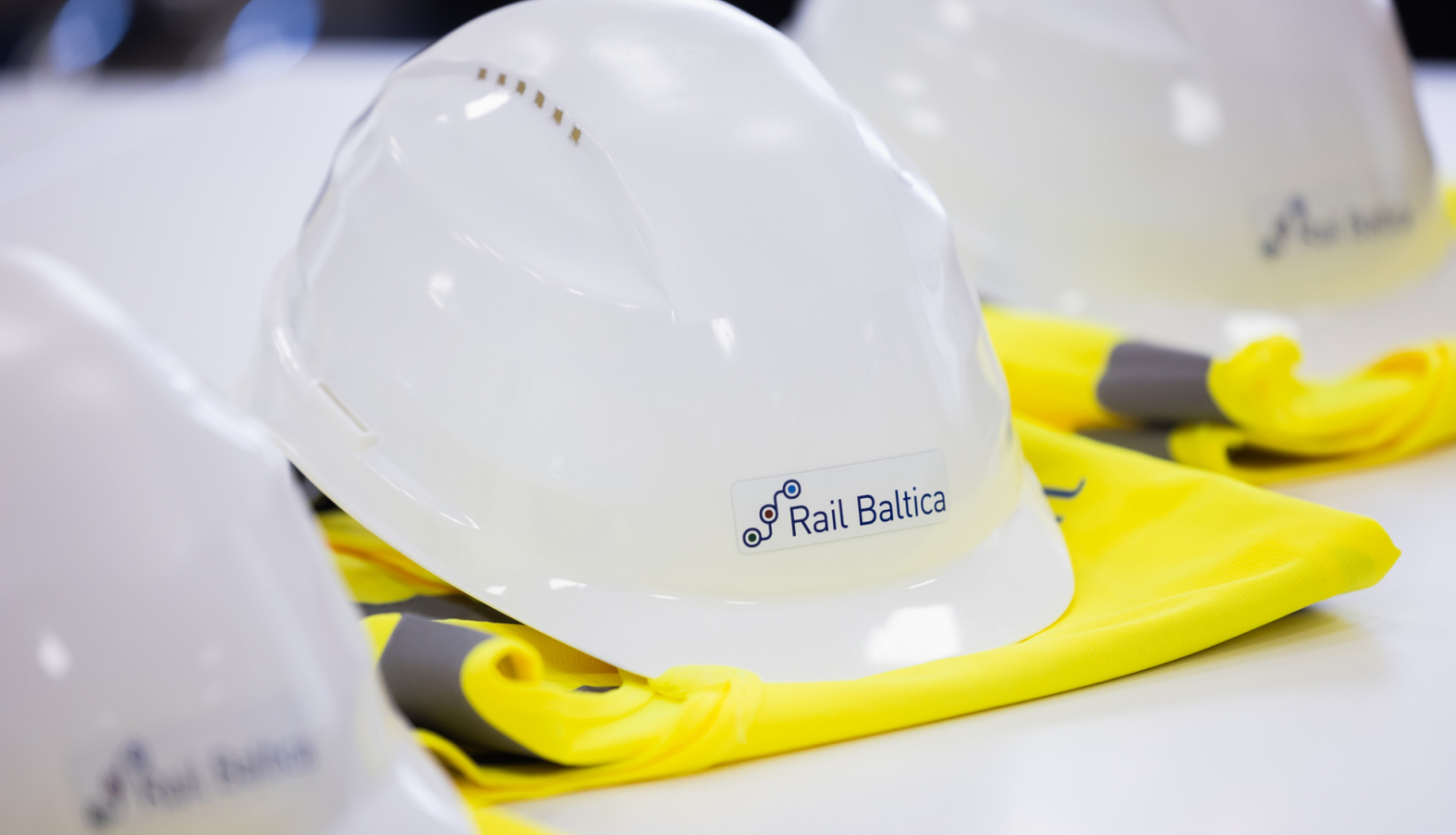 Uz galda novietoti balti aizsargķiveres ar uzlīmi 'Rail Baltica' un dzelteni atstarojoši vestes fragmenti